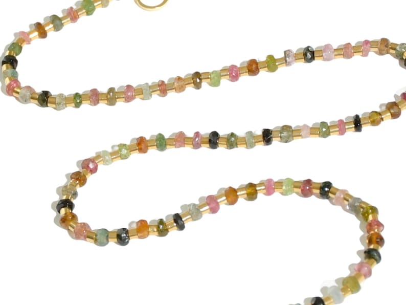 May include: A delicate gold chain necklace with small, multi-colored gemstones. The necklace features a variety of colors, including pink, green, and black.