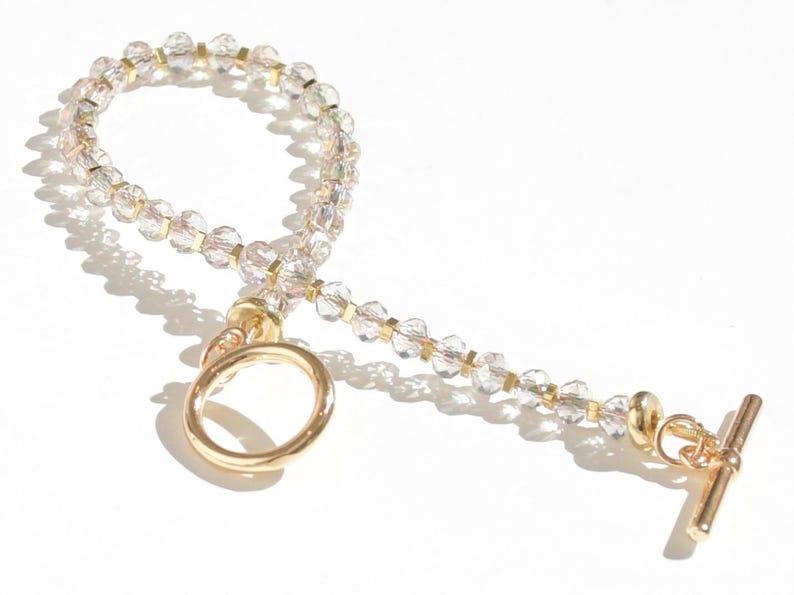 May include: A gold beaded bracelet with a clear crystal design. The bracelet has a gold toggle clasp and a gold ring.
