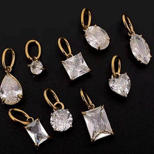 Gold diamond crystal charms displayed on a black background, each featuring a unique rhinestone cut — pear, heart, round, oval, marquise, princess, emerald, and radiant. The clear faceted stones shimmer in the light, framed in gold settings with looped bails for attaching to necklaces or bracelets. Elegant minimalist photo showing jewelry-making pendants available in both gold and silver options.  Perfect for diy jewelry to use as add-on charms for any charm necklace or charm bracelet.