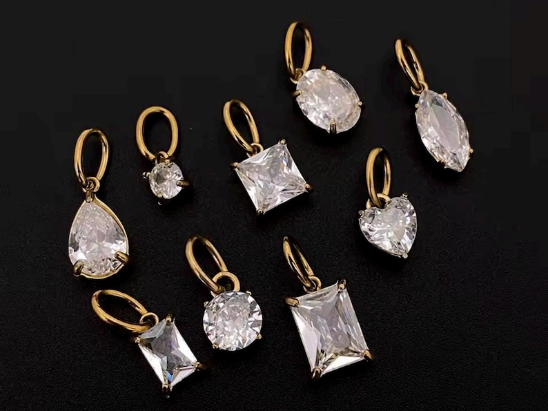 Gold diamond crystal charms displayed on a black background, each featuring a unique rhinestone cut — pear, heart, round, oval, marquise, princess, emerald, and radiant. The clear faceted stones shimmer in the light, framed in gold settings with looped bails for attaching to necklaces or bracelets. Elegant minimalist photo showing jewelry-making pendants available in both gold and silver options.  Perfect for diy jewelry to use as add-on charms for any charm necklace or charm bracelet.