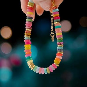May include: A colorful bracelet made of small, round, flat beads in various shades of pink, blue, green, yellow, and orange. The beads are strung on a gold chain.