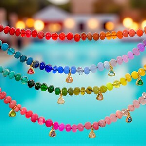 May include: A selection of colorful beaded necklaces in a rainbow of hues, held up against a blurred background. Each necklace features small, round beads and a gold-toned charm. The necklaces are in shades of red, orange, yellow, green, blue, and purple.