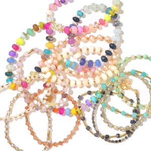 May include: A collection of colorful beaded bracelets with gold accents. The bracelets are made with various types of beads, including round, square, and oval shapes. The bracelets are arranged in a pile, with some overlapping.
