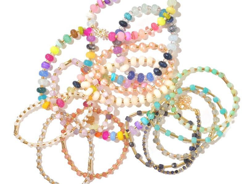 May include: A collection of colorful beaded bracelets with gold accents. The bracelets are made with various types of beads, including round, square, and oval shapes. The bracelets are arranged in a pile, with some overlapping.