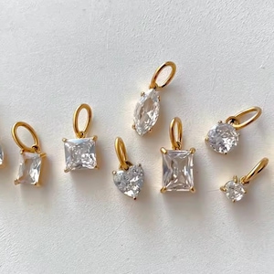 Gold diamond crystal charms displayed on a white background, each featuring a unique rhinestone cut — pear, heart, round, oval, marquise, princess, emerald, and radiant. The clear faceted stones shimmer in the light, framed in gold settings with looped bails for attaching to necklaces or bracelets. Elegant minimalist photo showing jewelry-making pendants available in both gold and silver options.  Perfect for diy jewelry to use as add-on charms for any charm necklace or charm bracelet.