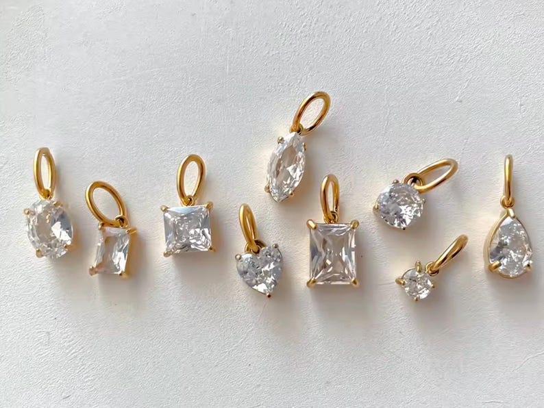 Gold diamond crystal charms displayed on a white background, each featuring a unique rhinestone cut — pear, heart, round, oval, marquise, princess, emerald, and radiant. The clear faceted stones shimmer in the light, framed in gold settings with looped bails for attaching to necklaces or bracelets. Elegant minimalist photo showing jewelry-making pendants available in both gold and silver options.  Perfect for diy jewelry to use as add-on charms for any charm necklace or charm bracelet.