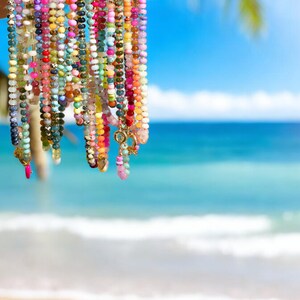 May include: A collection of colorful beaded necklaces with gold accents hanging against a blurred background of a beach and ocean.