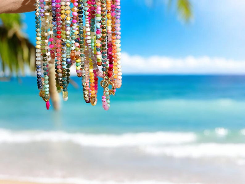 May include: A collection of colorful beaded necklaces with gold accents hanging against a blurred background of a beach and ocean.
