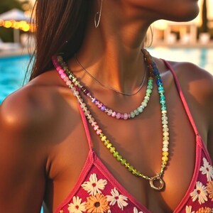 Multi-strand beaded necklace featuring a rainbow gradient of gemstone beads.  The necklace includes pink, purple, blue, green, and yellow beads, graduating smoothly from one color to the next.  The beads are faceted and appear to be semi-precious stones. The necklace has a gold clasp.  The necklace is layered, with one strand slightly shorter than the other. The style is bohemian and colorful.