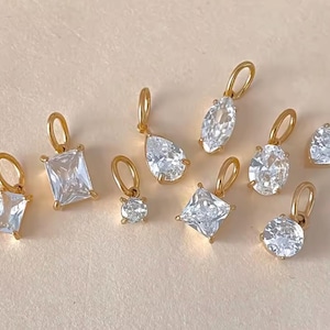 Gold diamond crystal charms displayed on a warm background, each featuring a unique rhinestone cut — pear, heart, round, oval, marquise, princess, emerald, and radiant. The clear faceted stones shimmer in the light, framed in gold settings with looped bails for attaching to necklaces or bracelets. Elegant minimalist photo showing jewelry-making pendants available in both gold and silver options.  Perfect for diy jewelry to use as add-on charms for any charm necklace or charm bracelet.