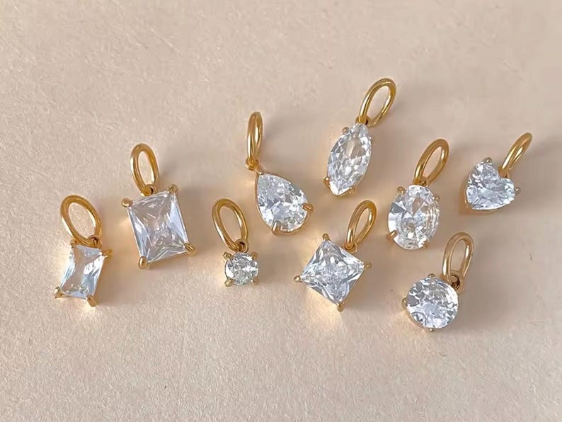 Gold diamond crystal charms displayed on a warm background, each featuring a unique rhinestone cut — pear, heart, round, oval, marquise, princess, emerald, and radiant. The clear faceted stones shimmer in the light, framed in gold settings with looped bails for attaching to necklaces or bracelets. Elegant minimalist photo showing jewelry-making pendants available in both gold and silver options.  Perfect for diy jewelry to use as add-on charms for any charm necklace or charm bracelet.
