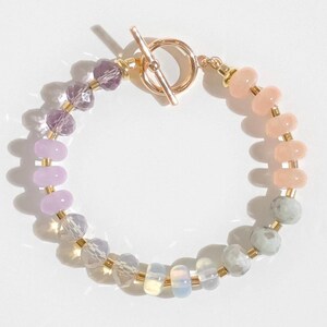 May include: A multi-colored beaded bracelet with a gold clasp. The bracelet features shades of purple, pink, and gray beads.