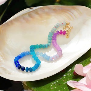 Hand-knotted gemstone necklace featuring turquoise, royal blue, and lilac beads with a gold clasp, laid out in a soft ombré gradient.