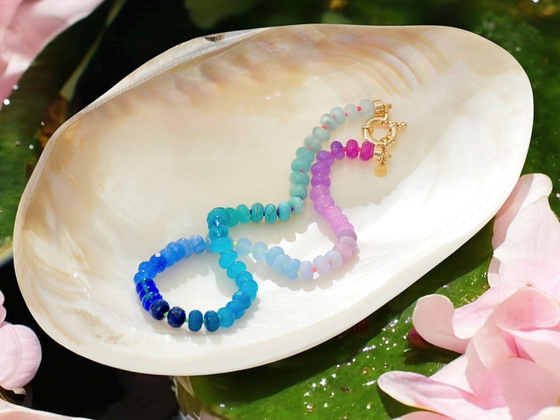 Hand-knotted gemstone necklace featuring turquoise, royal blue, and lilac beads with a gold clasp, laid out in a soft ombré gradient.