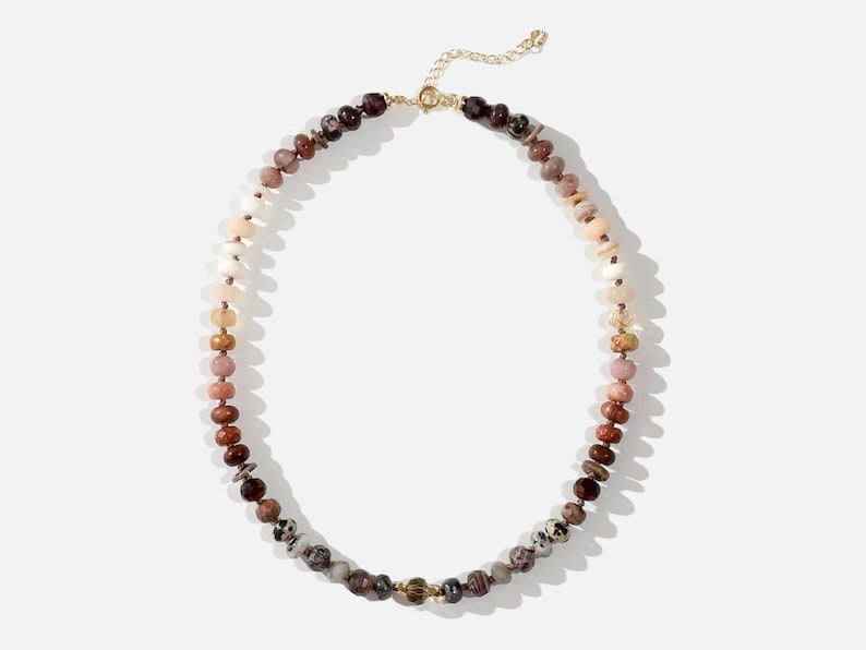 A long necklace made of various colored stones. The stones are in shades of brown, creams, white, and natural hues. The necklace has a gold clasp.