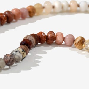 A long necklace made of various colored stones. The stones are in shades of brown, creams, white, and natural hues. The necklace has a gold clasp.