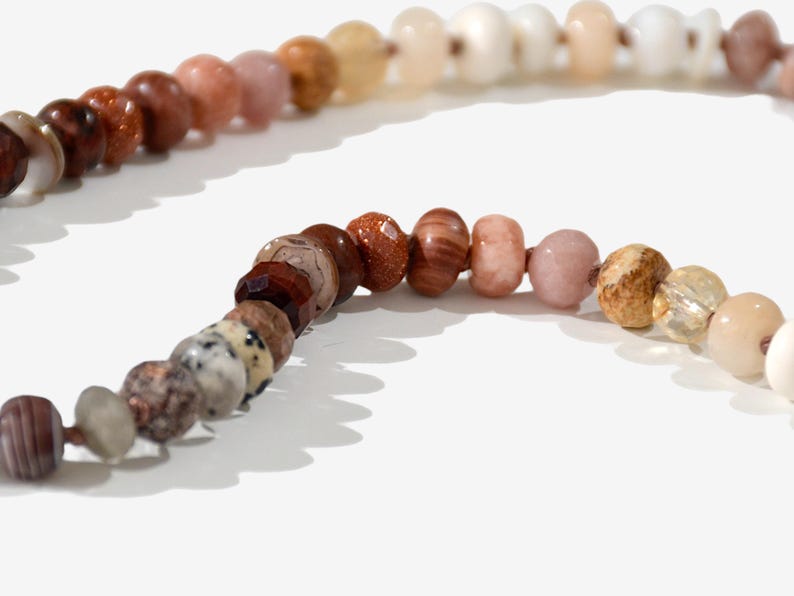 A long necklace made of various colored stones. The stones are in shades of brown, creams, white, and natural hues. The necklace has a gold clasp.