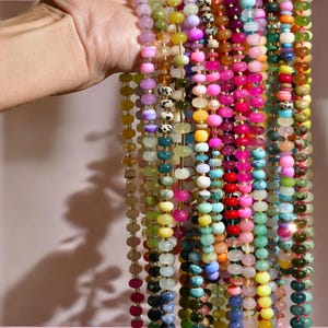 May include: A collection of colorful gemstone beads strung on multiple strands. The beads are various shapes and sizes, including round, oval, and irregular. The colors include pink, green, blue, yellow, and purple.