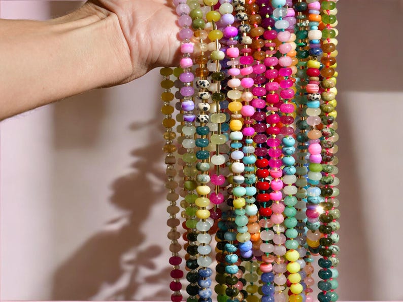 May include: A collection of colorful gemstone beads strung on multiple strands. The beads are various shapes and sizes, including round, oval, and irregular. The colors include pink, green, blue, yellow, and purple.