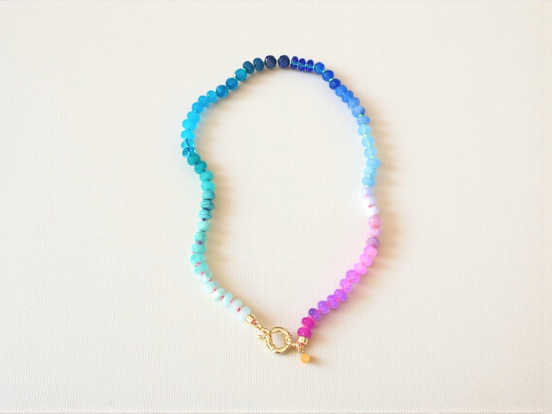 Gradient gemstone necklace featuring genuine pink, lavender, cobalt blue, and aqua blue faceted beads with 14k gold vermeil accents and a gold spring clasp on a beige surface.