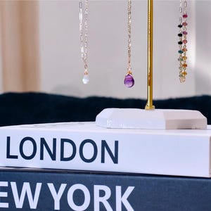 May include: Three gold necklaces with gemstone pendants displayed on a gold stand. The necklaces are on top of a white book with the words "LONDON" and "NEW YORK" printed on the front.
