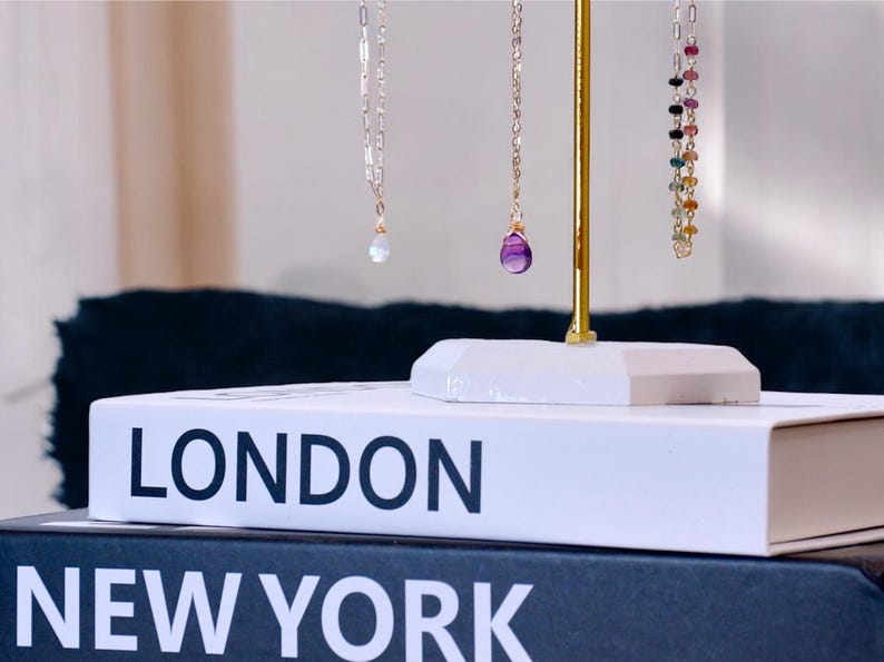 May include: Three gold necklaces with gemstone pendants displayed on a gold stand. The necklaces are on top of a white book with the words "LONDON" and "NEW YORK" printed on the front.