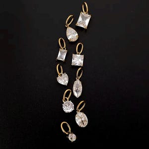 Gold diamond crystal charms displayed on a black background, each featuring a unique rhinestone cut — pear, heart, round, oval, marquise, princess, emerald, and radiant. The clear faceted stones shimmer in the light, framed in gold settings with looped bails for attaching to necklaces or bracelets. Elegant minimalist photo showing jewelry-making pendants available in both gold and silver options.  Perfect for diy jewelry to use as add-on charms for any charm necklace or charm bracelet.