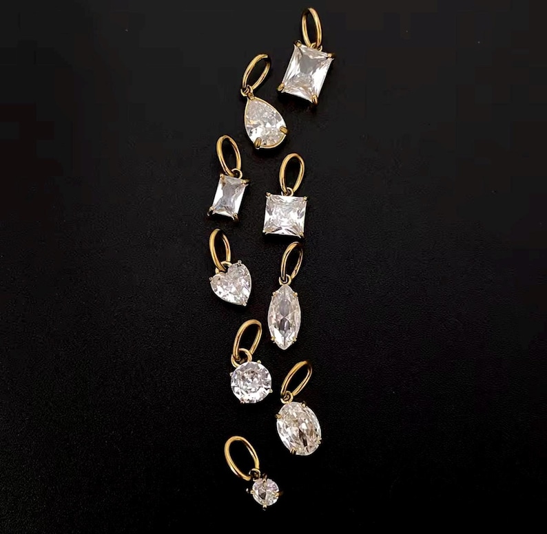 Gold diamond crystal charms displayed on a black background, each featuring a unique rhinestone cut — pear, heart, round, oval, marquise, princess, emerald, and radiant. The clear faceted stones shimmer in the light, framed in gold settings with looped bails for attaching to necklaces or bracelets. Elegant minimalist photo showing jewelry-making pendants available in both gold and silver options.  Perfect for diy jewelry to use as add-on charms for any charm necklace or charm bracelet.