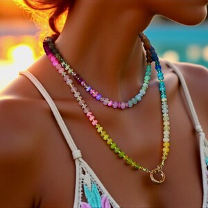 Multi-strand beaded necklace featuring a rainbow gradient of gemstone beads.  The necklace includes pink, purple, blue, green, and yellow beads, graduating smoothly from one color to the next.  The beads are faceted and appear to be semi-precious stones. The necklace has a gold clasp.  The necklace is layered, with one strand slightly shorter than the other. The style is bohemian and colorful.