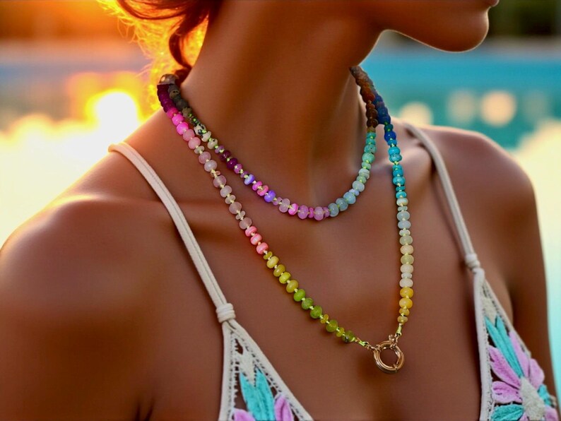 Multi-strand beaded necklace featuring a rainbow gradient of gemstone beads.  The necklace includes pink, purple, blue, green, and yellow beads, graduating smoothly from one color to the next.  The beads are faceted and appear to be semi-precious stones. The necklace has a gold clasp.  The necklace is layered, with one strand slightly shorter than the other. The style is bohemian and colorful.