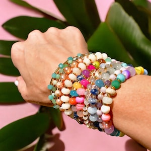 May include: A stack of colorful beaded bracelets with gold accents. The bracelets are made of various materials, including glass, stone, and wood. The bracelets are stacked on a person's wrist.