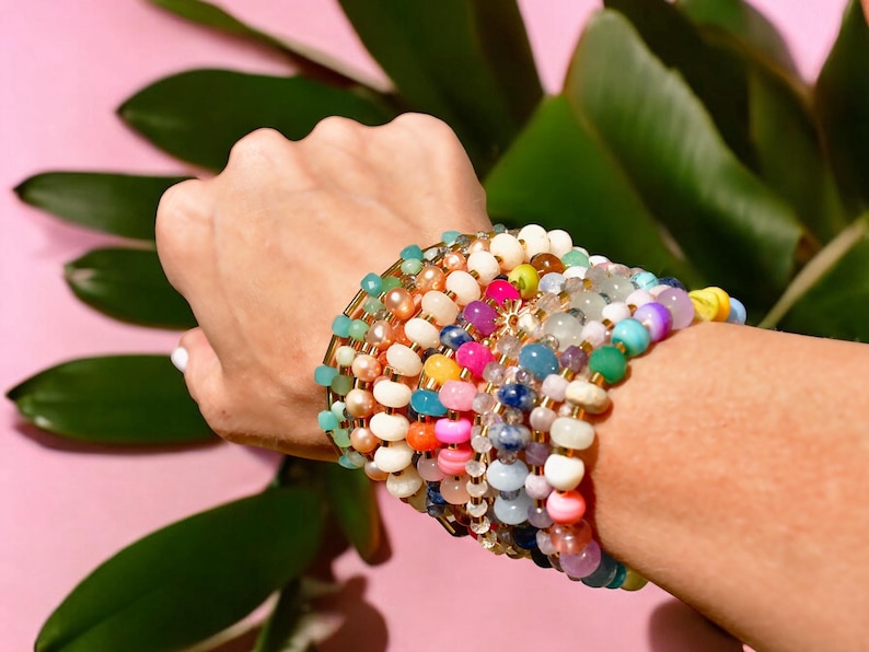 May include: A stack of colorful beaded bracelets with gold accents. The bracelets are made of various materials, including glass, stone, and wood. The bracelets are stacked on a person's wrist.