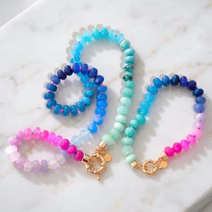 Gradient gemstone bracelet and matching necklace featuring genuine pink, lavender, cobalt blue, and aqua blue faceted beads with 14k gold vermeil accents and gold spring clasps on a white marble background.