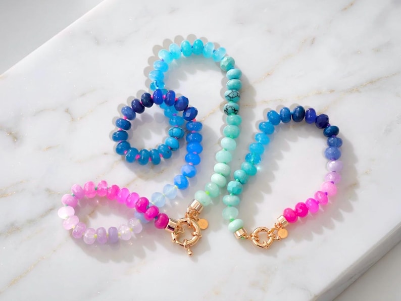 Gradient gemstone bracelet and matching necklace featuring genuine pink, lavender, cobalt blue, and aqua blue faceted beads with 14k gold vermeil accents and gold spring clasps on a white marble background.