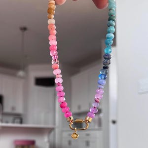 May include: A beaded necklace featuring a gradient of small, faceted round beads transitioning from warm tan and pink tones to cool blue and purple shades. The necklace includes a gold-toned oval carabiner clasp with a small, dangling flower charm.
