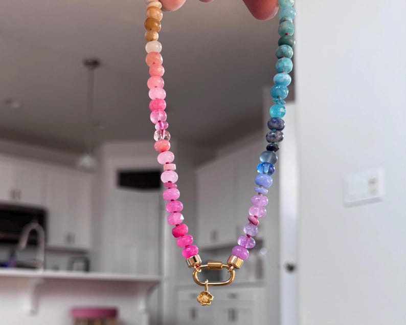 May include: A beaded necklace featuring a gradient of small, faceted round beads transitioning from warm tan and pink tones to cool blue and purple shades. The necklace includes a gold-toned oval carabiner clasp with a small, dangling flower charm.