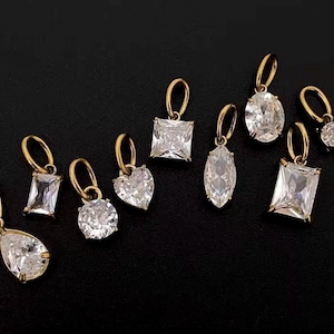 Gold diamond crystal charms displayed on a black background, each featuring a unique rhinestone cut — pear, heart, round, oval, marquise, princess, emerald, and radiant. The clear faceted stones shimmer in the light, framed in gold settings with looped bails for attaching to necklaces or bracelets. Elegant minimalist photo showing jewelry-making pendants available in both gold and silver options.  Perfect for diy jewelry to use as add-on charms for any charm necklace or charm bracelet.