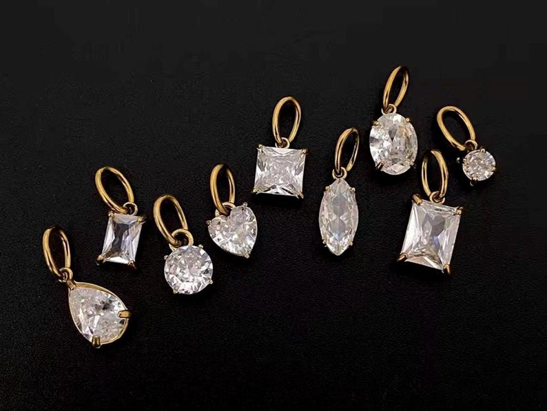 Gold diamond crystal charms displayed on a black background, each featuring a unique rhinestone cut — pear, heart, round, oval, marquise, princess, emerald, and radiant. The clear faceted stones shimmer in the light, framed in gold settings with looped bails for attaching to necklaces or bracelets. Elegant minimalist photo showing jewelry-making pendants available in both gold and silver options.  Perfect for diy jewelry to use as add-on charms for any charm necklace or charm bracelet.