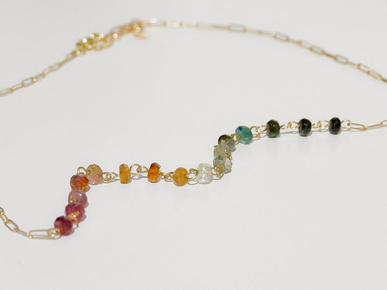 May include: A gold chain necklace with a rainbow of gemstone beads. The beads are in a zig-zag pattern and include pink, orange, yellow, white, green, and black.