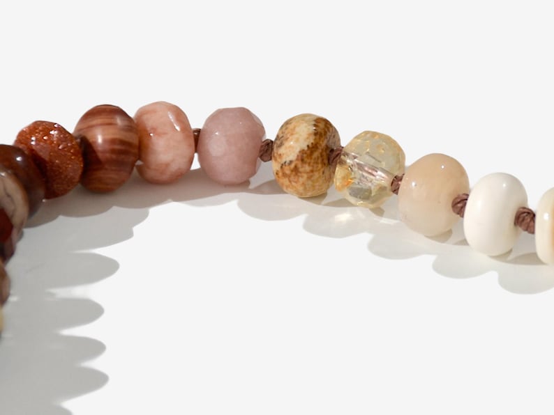 A long necklace made of various colored stones. The stones are in shades of brown, creams, white, and natural hues. The necklace has a gold clasp.
