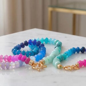 Gradient gemstone bracelet featuring genuine pink, lavender, cobalt blue, and aqua blue faceted beads with 14k gold vermeil accents and a gold toggle clasp on a white marble background.