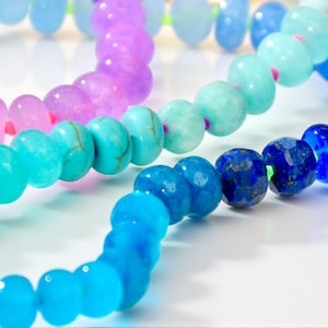 Hand-knotted gemstone necklace featuring turquoise, royal blue, and lilac beads with a gold clasp, laid out in a soft ombré gradient.