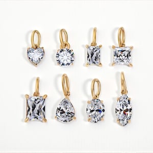 Gold diamond crystal charms displayed on a white background, each featuring a unique rhinestone cut — pear, heart, round, oval, marquise, princess, emerald, and radiant. The clear faceted stones shimmer in the light, framed in gold settings with looped bails for attaching to necklaces or bracelets. Elegant minimalist photo showing jewelry-making pendants available in both gold and silver options.  Perfect for diy jewelry to use as add-on charms for any charm necklace or charm bracelet.