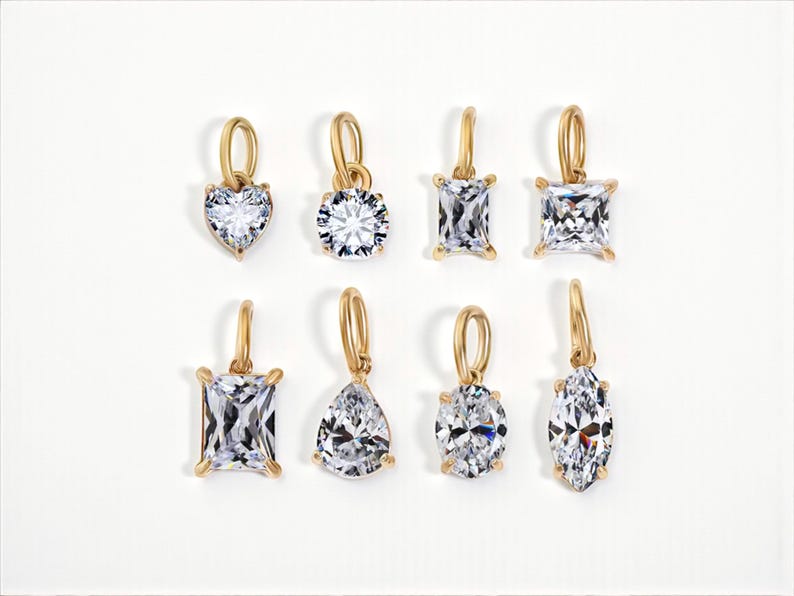 Gold diamond crystal charms displayed on a white background, each featuring a unique rhinestone cut — pear, heart, round, oval, marquise, princess, emerald, and radiant. The clear faceted stones shimmer in the light, framed in gold settings with looped bails for attaching to necklaces or bracelets. Elegant minimalist photo showing jewelry-making pendants available in both gold and silver options.  Perfect for diy jewelry to use as add-on charms for any charm necklace or charm bracelet.