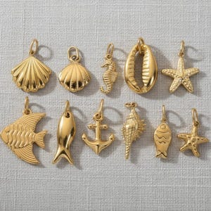 Peut inclure: Une collection de breloques nautiques dorées, comprenant des coquillages, des hippocampes, des étoiles de mer, des poissons et une ancre. Ces pendentifs sont conçus avec des détails complexes et sont présentés sur une surface en lin neutre.