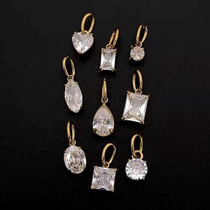 Gold diamond crystal charms displayed on a black background, each featuring a unique rhinestone cut — pear, heart, round, oval, marquise, princess, emerald, and radiant. The clear faceted stones shimmer in the light, framed in gold settings with looped bails for attaching to necklaces or bracelets. Elegant minimalist photo showing jewelry-making pendants available in both gold and silver options.  Perfect for diy jewelry to use as add-on charms for any charm necklace or charm bracelet.
