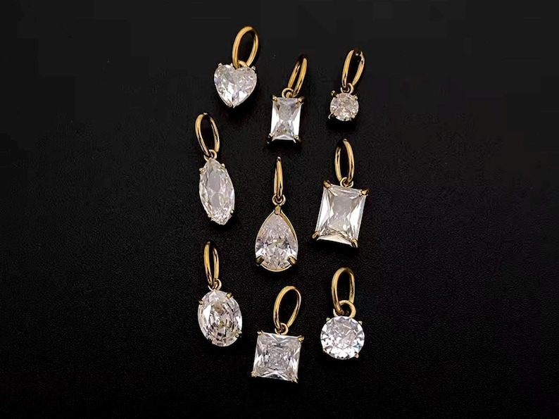 Gold diamond crystal charms displayed on a black background, each featuring a unique rhinestone cut — pear, heart, round, oval, marquise, princess, emerald, and radiant. The clear faceted stones shimmer in the light, framed in gold settings with looped bails for attaching to necklaces or bracelets. Elegant minimalist photo showing jewelry-making pendants available in both gold and silver options.  Perfect for diy jewelry to use as add-on charms for any charm necklace or charm bracelet.