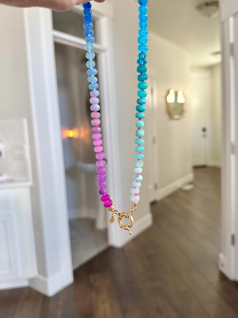 Hand-knotted gemstone necklace featuring turquoise, royal blue, and lilac beads with a gold clasp, laid out in a soft ombré gradient.