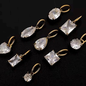 Gold diamond crystal charms displayed on a black background, each featuring a unique rhinestone cut — pear, heart, round, oval, marquise, princess, emerald, and radiant. The clear faceted stones shimmer in the light, framed in gold settings with looped bails for attaching to necklaces or bracelets. Elegant minimalist photo showing jewelry-making pendants available in both gold and silver options.  Perfect for diy jewelry to use as add-on charms for any charm necklace or charm bracelet.