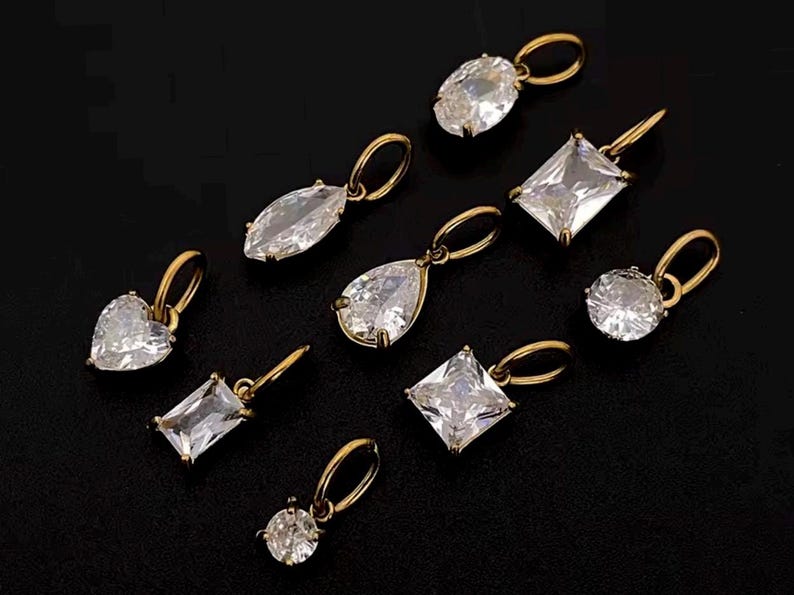 Gold diamond crystal charms displayed on a black background, each featuring a unique rhinestone cut — pear, heart, round, oval, marquise, princess, emerald, and radiant. The clear faceted stones shimmer in the light, framed in gold settings with looped bails for attaching to necklaces or bracelets. Elegant minimalist photo showing jewelry-making pendants available in both gold and silver options.  Perfect for diy jewelry to use as add-on charms for any charm necklace or charm bracelet.